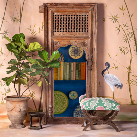 Decorative setting with a large mirror, plant, and stool against a wall with bamboo art.
